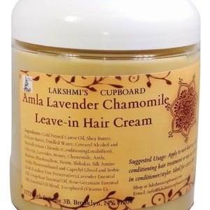 May include: A glass jar with a white lid containing a light yellow cream. The label is orange and brown with the text "LAKSHMI'S CUPBOARD Amla Lavender Chamomile Leave-in Hair Cream". The ingredients and suggested usage are listed on the label.