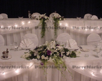 May include: A decorated wedding reception table with white linens, chairs, and floral arrangements. The table is adorned with white and purple flowers, greenery, and string lights, creating an elegant ambiance for a special event.