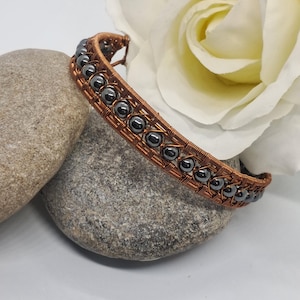 May include: A handcrafted copper wire bracelet with rows of dark gray beads. The bracelet is displayed on a gray stone, with a white rose in the background. The bracelet is a unique piece of jewelry.