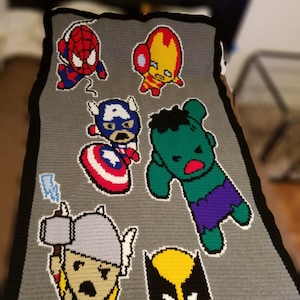 Op de afbeelding: Een gehaakte deken met een kleurrijk scala aan superheldenfiguren, waaronder Spider-Man, Iron Man, Captain America, Hulk, Thor en Wolverine. De personages zijn in cartoon-stijl afgebeeld en zijn geplaatst op een grijze achtergrond.