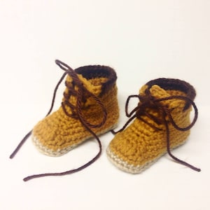 Puede incluir: Un par de botines de bebé tejidos a crochet en color amarillo mostaza con cordones marrones. Los botines tienen un ribete blanco alrededor de la parte inferior.
