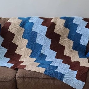 Puede incluir: Una manta de crochet con un patrón de chevron en tonos marrones, azules y beige. La manta está colocada sobre un sofá marrón.