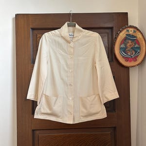 May include: A cream-colored button-down shirt with a high collar, three-quarter length sleeves, and two square patch pockets on the front. The shirt features a central vertical row of white buttons and a visible brand label at the inner neckline.