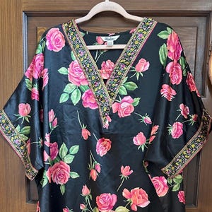 May include: A black V-neck tunic featuring a vibrant pink rose and green leaf floral print. The garment includes a decorative multi-colored floral trim along the neckline and sleeve cuffs. A white label with the text Winlar is visible at the neck.