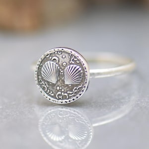 Peut inclure: Bague en argent avec un motif circulaire orné de deux coquillages. La bague présente une surface texturée et détaillée, ainsi qu'un anneau lisse. La bague est présentée sur une surface réfléchissante.