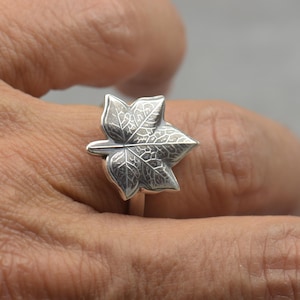 Puede incluir: Un anillo de plata con un diseño detallado de hoja. La hoja del anillo tiene vetas intrincadas y está engastada en una banda delgada. El anillo se muestra en una mano, destacando la delicada artesanía de la joya.