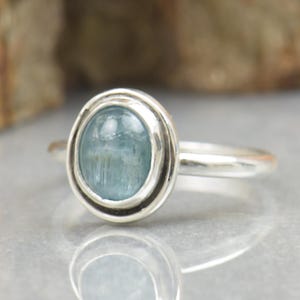 Könnte beinhalten: Ein silberner Ring mit einem hellblauen ovalen Edelstein, der in einer Zargenfassung gefasst ist. Der Ring liegt auf einer weißen Oberfläche.