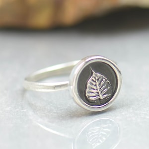 May include: A silver ring with a black circular design featuring a raised silver leaf.