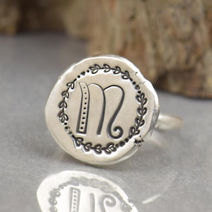 Könnte beinhalten: Silberner Ring mit einem kreisförmigen Gesicht, das den Buchstaben "M" in einer stilisierten Schriftart zeigt, umgeben von einem Lorbeerkranz-Design. Der Ring ist vor einem neutralen Hintergrund platziert, mit einer Reflexion des Rings darunter.