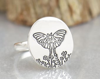 May include: A silver ring featuring a flat, oval-shaped face engraved with a detailed moth design positioned above a cluster of small flowers. The metal surface is polished and reflective, showcasing the dark, etched lines of the insect.