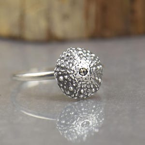 Puede incluir: Un anillo de plata con un punto focal en forma de concha de erizo de mar esférica y texturizada. La superficie metálica tiene un acabado oxidado oscuro en los detalles complejos y abultados de la concha, contrastando con una banda lisa.