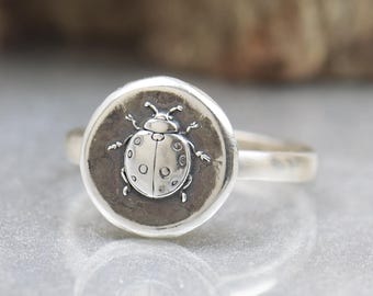 May include: A silver ring featuring a detailed ladybug design. The ladybug is centered on a circular, textured background. The ring's band is smooth and polished, reflecting the light. The ring is set against a blurred background.
