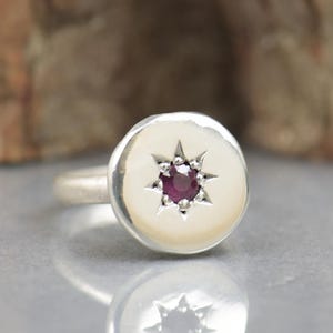 May include: A silver ring with a round, white, textured surface featuring a small, dark red gemstone in the centre of a starburst design.
