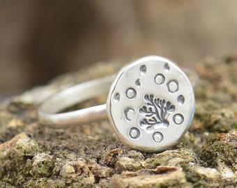Puede incluir: Un anillo de plata con una cara redonda que presenta un diseño de árbol y un patrón circular. El anillo está sobre un trozo de madera.