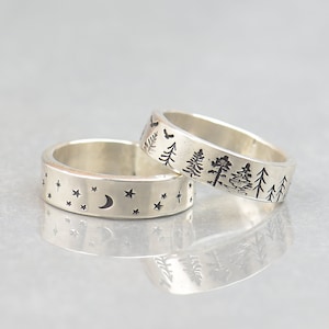 May include: Two silver rings with etched designs. One ring features a crescent moon and stars, the other depicts a forest scene with trees and a flying bird. The rings are displayed on a reflective surface.