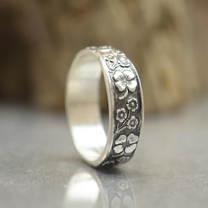 Könnte beinhalten: Ein silberner Ring mit floralem Muster. Der Ring hat ein sich wiederholendes Muster aus Blumen und Blättern um das Band. Der Ring wird auf einer reflektierenden Oberfläche gezeigt.