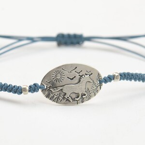 May include: A blue cord bracelet with a silver oval charm featuring a stylized image of a horse running through a mountain landscape with a sun and birds in the sky.