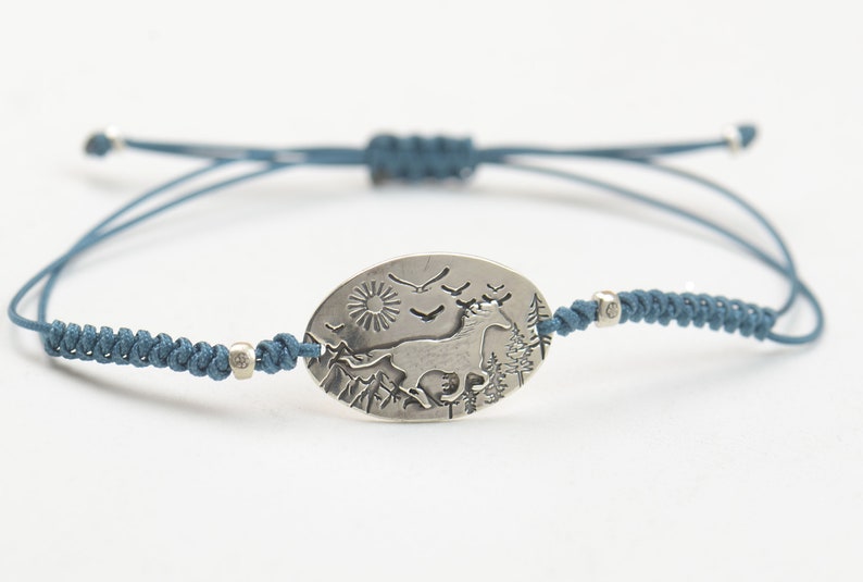 May include: A blue cord bracelet with a silver oval charm featuring a stylized image of a horse running through a mountain landscape with a sun and birds in the sky.