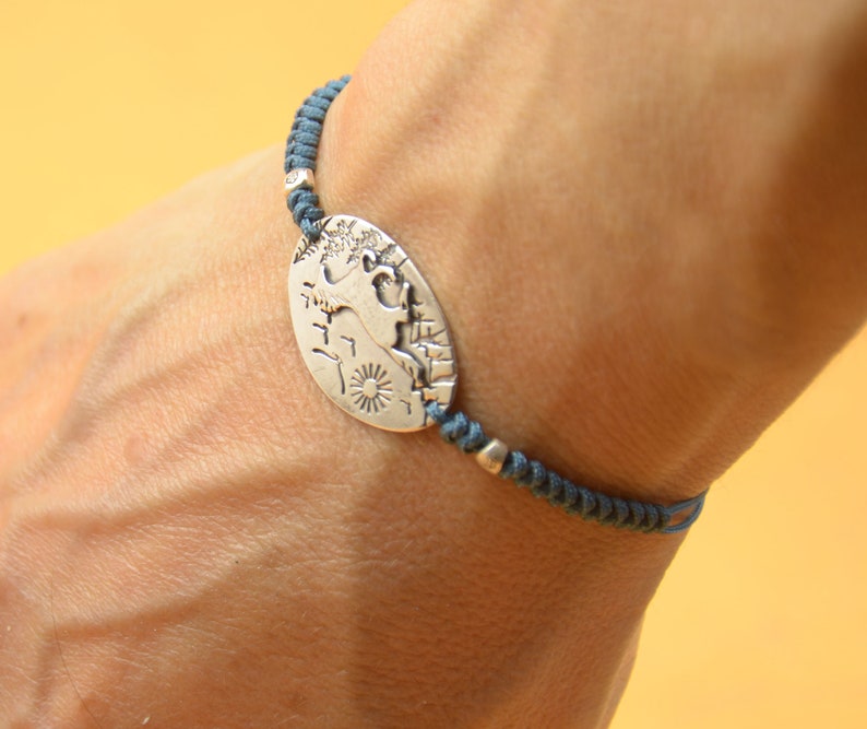 May include: A silver charm bracelet with a detailed design of a mythical creature on a round charm. The bracelet is made of dark blue cord.