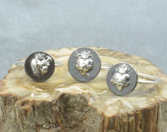 May include: Three silver rings with a raised image of a heart pierced by a sword and surrounded by a crown of thorns. The rings are displayed on a piece of wood.