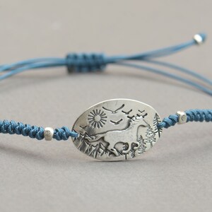 May include: A blue cord bracelet with a silver oval charm featuring a stylized image of a horse running through a forest with a sun and birds in the sky.