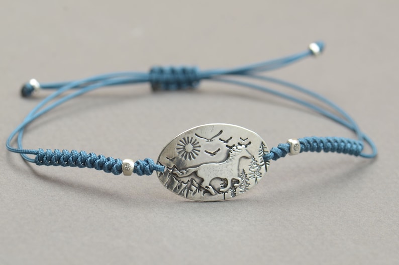 May include: A blue cord bracelet with a silver oval charm featuring a stylized image of a horse running through a forest with a sun and birds in the sky.