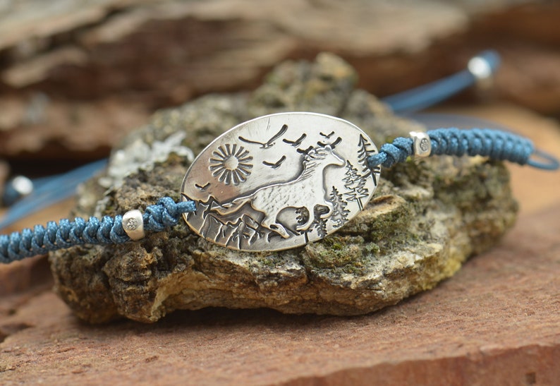 May include: A silver bracelet with a blue cord and a silver charm featuring a deer running through a forest scene with a sun and birds.