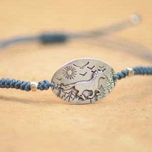 May include: A blue cord bracelet with a silver oval charm featuring a running horse, a sun, birds, and trees.