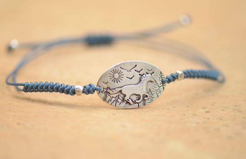 May include: A blue cord bracelet with a silver oval charm featuring a running horse, a sun, birds, and trees.