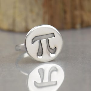 Puede incluir: Un anillo de plata con una cara circular que presenta el símbolo matemático pi grabado en relieve y en tono oscuro en el centro. El metal tiene un acabado liso y pulido, y el diseño está centrado en la parte superior redonda de la banda.