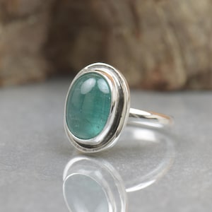 Può includere: Un anello in argento con una grande pietra preziosa ovale color verde acqua. La pietra è incastonata in una lunetta con un design a doppia fascia. L'anello ha una finitura lucida. La gemma misura circa 1,5 cm di lunghezza e 1 cm di larghezza.
