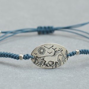 May include: A blue cord bracelet with a silver oval charm featuring an engraved image of a horse running through a mountain landscape with a sun and birds in the sky.