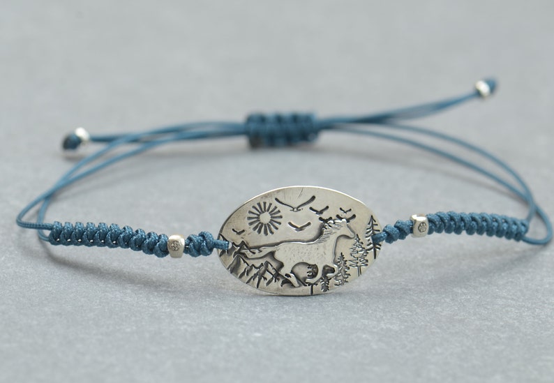 May include: A blue cord bracelet with a silver oval charm featuring an engraved image of a horse running through a mountain landscape with a sun and birds in the sky.