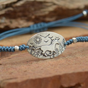 May include: A blue cord bracelet with a silver oval charm featuring a running horse, a sun, and birds in flight against a mountain backdrop.