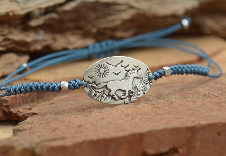 May include: A blue cord bracelet with a silver oval charm featuring a running horse, a sun, and birds in flight against a mountain backdrop.
