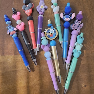 May include: A collection of colorful pens with decorative toppers featuring cartoon characters. The pens have various colors, including blue, black, red, and green. The toppers include Mickey Mouse, Stitch, and Care Bears.