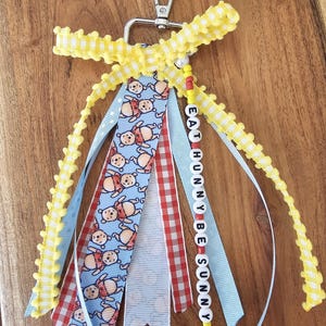 May include: A handmade keychain with a silver clasp, yellow beaded trim, and colorful ribbons. The ribbons feature Winnie the Pooh, red gingham, and the text "EAT HUNNY BE SUNNY" on letter beads. A cheerful and whimsical accessory.