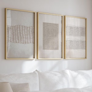 以下が含まれることがあります： 白い背景と茶色の線が入った、金色の額縁に入った抽象的なアートプリントが3枚。プリントは白い壁に、白いソファの上に白いクッションを置いて飾られています。ソファは、木のコーヒーテーブルと敷物のあるリビングルームにあります。