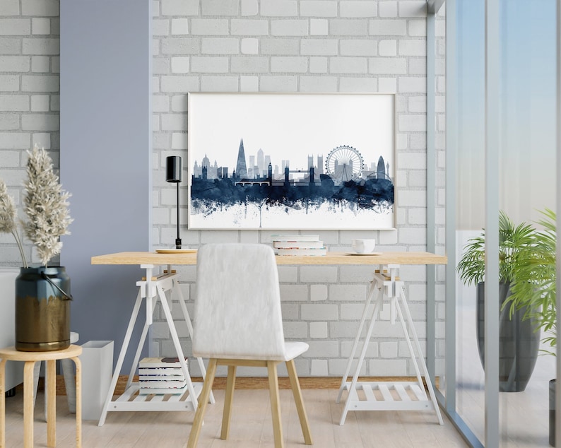 May include: A white desk with a wooden top and white trestle legs. A white chair sits in front of the desk. A framed print of a blue watercolor cityscape of London hangs on a white brick wall behind the desk. A potted plant sits on the floor next to a window.