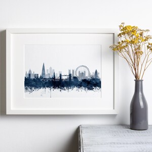 May include: A framed print of a watercolor cityscape of London, England. The skyline is depicted in shades of blue, with iconic landmarks like the London Eye, Big Ben, and Tower Bridge visible. The print is set against a white background and framed in a white frame.