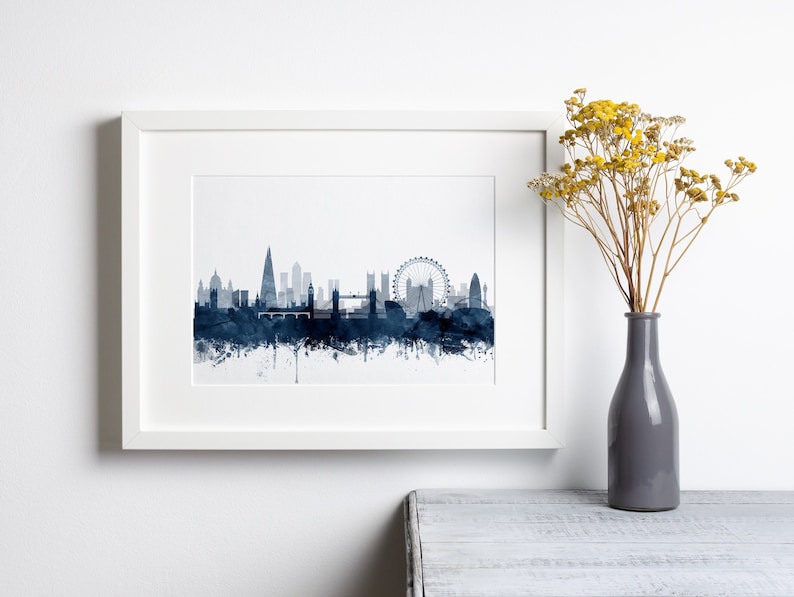 May include: A framed print of a watercolor cityscape of London, England. The skyline is depicted in shades of blue, with iconic landmarks like the London Eye, Big Ben, and Tower Bridge visible. The print is set against a white background and framed in a white frame.