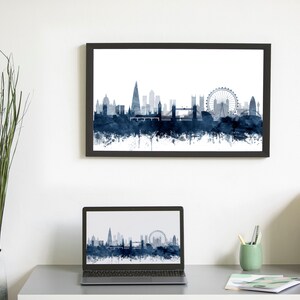 May include: A framed print of a watercolor cityscape of London, England, with the London Eye and other iconic landmarks in blue and white. A laptop computer with the same image on the screen is on a desk with a vase of greenery, a cup of pencils, and a magazine holder.