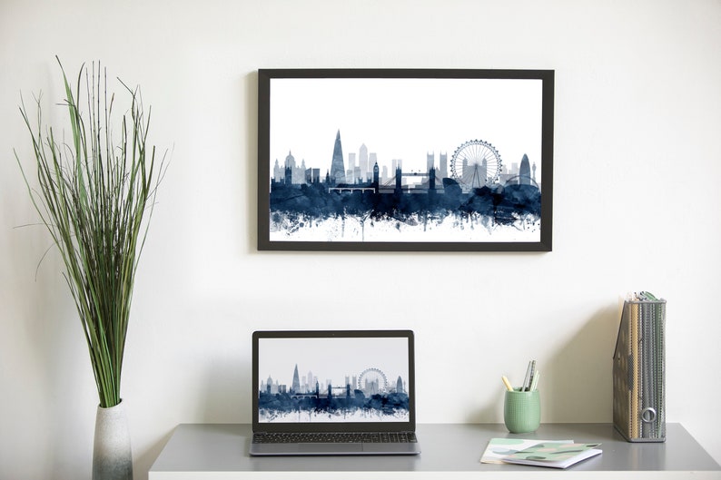 May include: A framed print of a watercolor cityscape of London, England, with the London Eye and other iconic landmarks in blue and white. A laptop computer with the same image on the screen is on a desk with a vase of greenery, a cup of pencils, and a magazine holder.