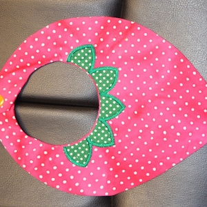 May include: Pink and white polka dot baby bib with green and white polka dot leaves around the neck opening. The bib has a yellow button at the top.