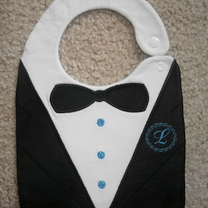 May include: A black and white bib with a tuxedo design, featuring a bow tie, a white shirt, and three blue buttons. The bib has a blue embroidered letter "L" in a circle on the right side.