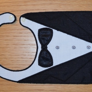 May include: A black and white bib with an embroidered tuxedo design. The bib has a white collar, a black bow tie, and three white buttons.