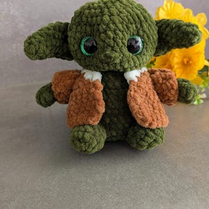 K&ouml;nnte beinhalten: Handgemachte gr&uuml;ne H&auml;kelpuppe mit gro&szlig;en, glitzernden Augen und gro&szlig;en Ohren. Die Puppe tr&auml;gt eine braune Jacke mit wei&szlig;em Besatz. Die Puppe ist eine Figur aus einem beliebten Science-Fiction-Franchise. Gelbe Blumen sind im Hintergrund.