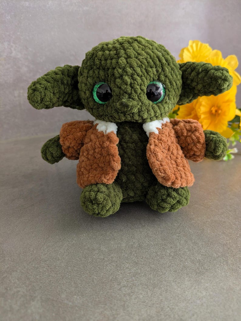 K&ouml;nnte beinhalten: Handgemachte gr&uuml;ne H&auml;kelpuppe mit gro&szlig;en, glitzernden Augen und gro&szlig;en Ohren. Die Puppe tr&auml;gt eine braune Jacke mit wei&szlig;em Besatz. Die Puppe ist eine Figur aus einem beliebten Science-Fiction-Franchise. Gelbe Blumen sind im Hintergrund.