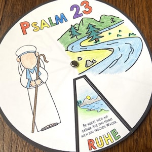 May include: A circular, illustrated craft featuring Psalm 23. The design includes a shepherd, a river scene, and the word "RUHE". The text "PSALM 23" is at the top. The craft is on a wooden surface.