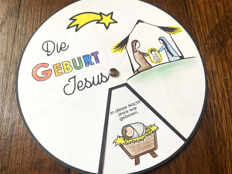 K&ouml;nnte beinhalten: Ein kreisf&ouml;rmiges, illustriertes Weihnachtsbastelwerk mit den Worten "Die Geburt Jesus" in bunten Buchstaben. Das Bild zeigt die Weihnachtsgeschichte mit Maria, Josef und Jesuskind und einen Sternschnuppen.